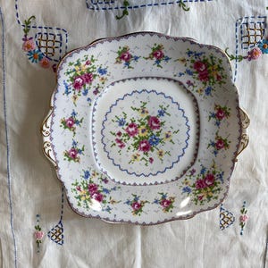 May include: A white square serving plate with a floral design. The plate has a scalloped edge and a gold rim. The floral design is in shades of pink, blue, and yellow.