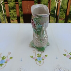 May include: A green and white ceramic vase with a floral design. The vase has a pearlescent glaze and is shaped like a boot.