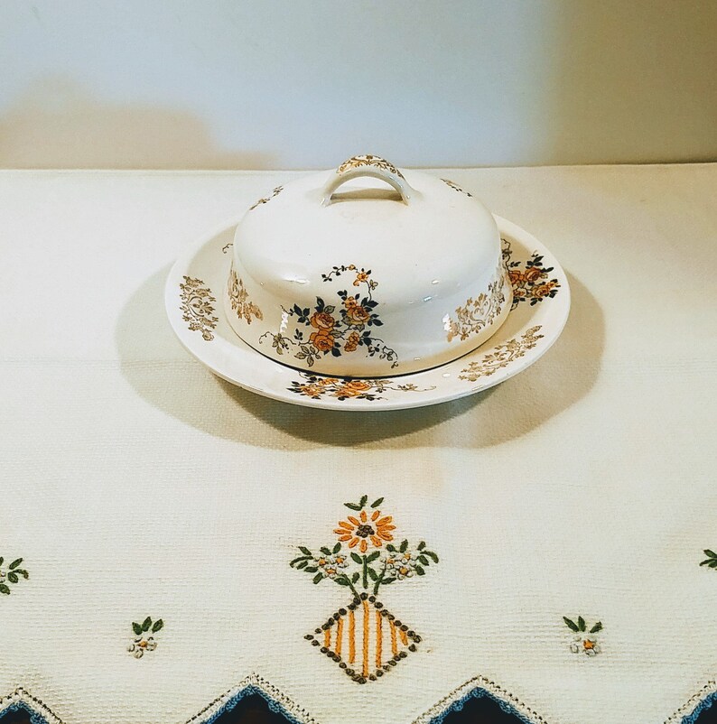 Puede incluir: Un mantequillero de cer&aacute;mica blanca con un dise&ntilde;o floral. El mantequillero tiene una tapa y una base con un borde festoneado. El mantequillero est&aacute; sobre un mantel blanco con flores bordadas.