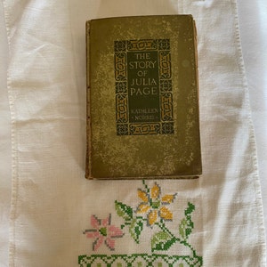 Puede incluir: Un libro vintage titulado "The Story of Julia Page" de Kathleen Norris. El libro tiene una cubierta verde con letras doradas y diseños intrincados. El libro está sobre un mantel de lino blanco con un diseño floral bordado en verde y amarillo.