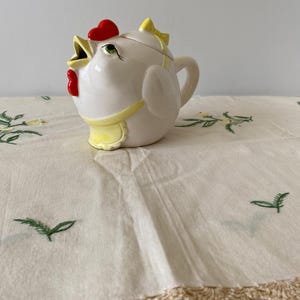 Könnte beinhalten: Ein weißes Keramik-Milchkännchen in Form eines Huhns mit rotem Kamm und Kehllappen, einer gelben Schürze und einer gelben Schleife auf dem Deckel. Das Huhn hat einen weit geöffneten Schnabel, grüne Augen und einen geschwungenen Henkel.
