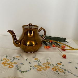 Peut inclure: Une théière de couleur cuivre avec une finition brillante repose sur une nappe blanche avec des fleurs brodées. La théière a un corps rond et un bec courbé.