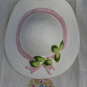 May include: A white ceramic hat with a pink ribbon and green leaves. The hat is decorated with a woven pattern and has a raised rim.