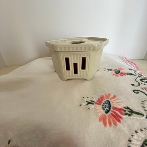 May include: A white ceramic tea warmer with a hexagonal base and a decorative design. The warmer has a small opening in the center and four cut-out sections on the sides.