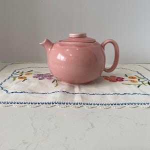 Puede incluir: Una tetera de cerámica rosa con tapa y asa a juego. La tetera está sobre un mantel individual bordado blanco con diseños florales en tonos morados, naranjas y amarillos. El mantel tiene un borde azul.