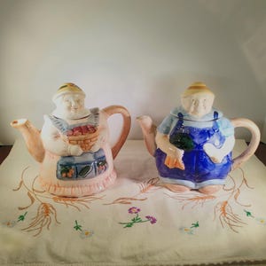 Può includere: Due teiere in ceramica a forma di persone, una rosa e una blu, con dettagli dipinti di un contadino e una contadina. Le teiere sono su una tovaglia bianca con grano e fiori ricamati.