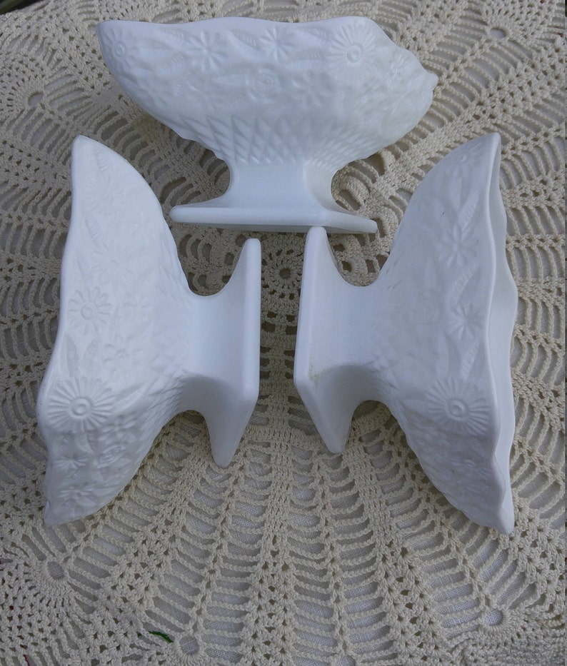 May include: Set of three white milk glass bud vases with a floral design. The vases are in the shape of a bowl with a wide base.
