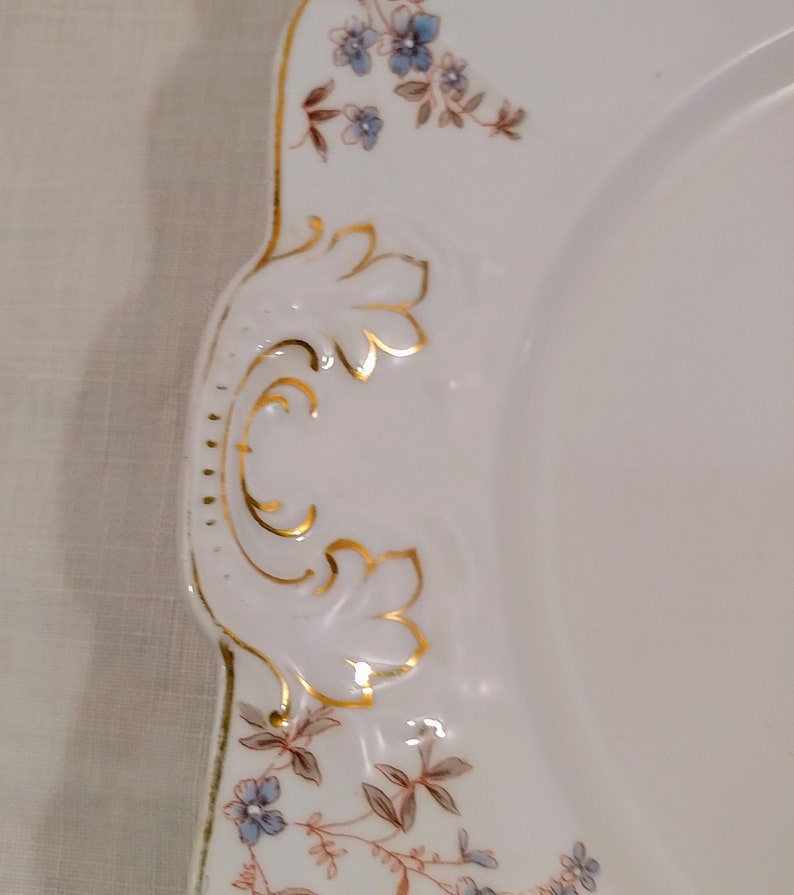 May include: White porcelain plate with a gold rim and floral design. The plate has a scalloped edge and a decorative handle.