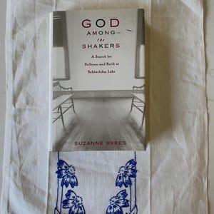 May include: A white book with the title "God Among the Shakers: A Search for Stillness and Faith at Sabbathday Lake" by Suzanne Skees. The book cover features a black and white photograph of a room with two beds.