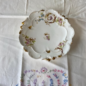 May include: A white porcelain serving plate with a scalloped edge and gold trim. The plate features floral designs in shades of pink, purple, blue, and green. The plate is placed on a white embroidered tablecloth.