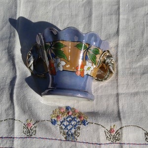 May include: A blue and orange ceramic planter with floral designs and two elephant-shaped handles. The planter is sitting on a white embroidered tablecloth.