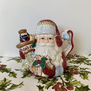 Puede incluir: Una tetera de cerámica de Papá Noel con gorro, barba y traje rojos y blancos. La tetera presenta un diseño detallado con un cascanueces y regalos. El asa tiene un diseño a rayas rojas y blancas. La tetera está sobre un mantel blanco con patrones de acebo verde y bayas rojas.