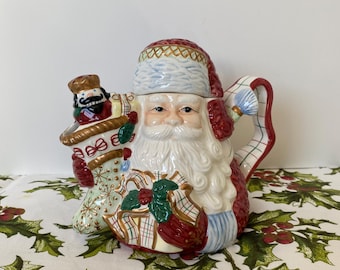 May include: A ceramic Santa Claus teapot with a red and white hat, beard, and suit. The teapot features a detailed design with a nutcracker and gifts. The handle is a red and white striped design. The teapot is on a white tablecloth with green holly and red berry patterns.