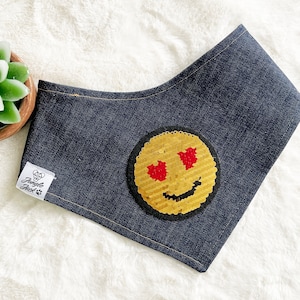 May include: A dark blue denim dog bandana featuring a sequined emoji face. The emoji has gold sequins for the face, red heart eyes, and a black stitched outline. A white tag with the text "Jungle Jack" is sewn on the side.