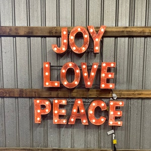 May include: Red metal letters spelling out "JOY LOVE PEACE" with light bulbs in each letter. The letters are mounted on a wall with a wood plank behind them.