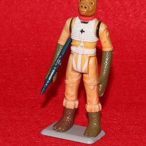May include: A vintage action figure of a reptilian alien character. The figure is dressed in an orange jumpsuit with a white vest and black accents. It holds a blue blaster and stands on a gray base. The figure has a brown head and green textured arms and boots.