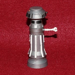 May include: A silver and gray robot figure with a white arm extending to the right. The robot has a cylindrical body with vertical white accents and a rounded top. The figure is standing on a gray base.