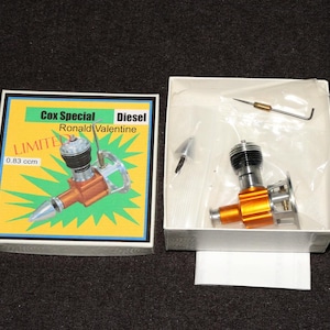 May include: A Cox Special Ronald Valentine 0.83 ccm diesel engine in a white box with a matching illustrated lid. The engine features an orange metallic body, a silver propeller hub, and a black cooling head. Included are small metal tools in plastic bags.