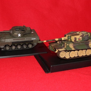 May include: Two model tanks on black display stands against a red background. One tank is olive green with a white star, the other is camouflage with brown, green, and tan. The tanks are detailed with tracks and turrets.