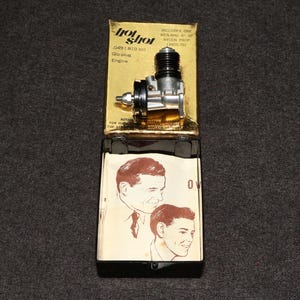 May include: A Hot Shot .049 (.819 cc) Glo-plug engine in a gold box with black text, including a Wen-Mac 6 inch 4-blade nylon prop. The black plastic base contains a paper manual featuring a brown-toned illustration of two smiling individuals.