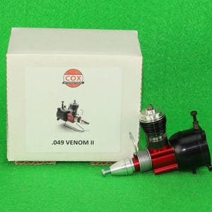 May include: A Cox International .049 Venom II model engine with a red and black body and silver components, positioned next to a white cardboard box featuring the Cox logo and the text .049 VENOM II printed on the front label.