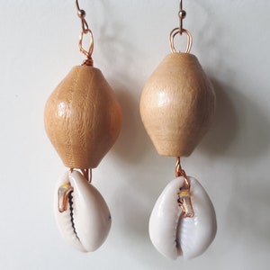 May include: A pair of handmade earrings featuring light brown wooden beads and white cowrie shells. The beads are a rounded, elongated shape, and the shells are attached with copper wire. The earrings have copper ear wires.