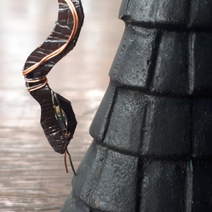 May include: Close-up of a dark brown, snake-like sculpture with copper wire detailing. The sculpture has a textured surface and is partially attached to a black, tiered structure. The sculpture's head is visible.