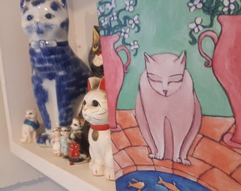 May include: A collection of cat-themed decor on a white shelf. Items include ceramic cat figurines in blue and white, white with red collar, and a painting of a pink cat with fish in a pond. The painting has pink vases with white flowers.