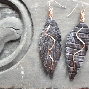 May include: A pair of handmade earrings featuring dark gray, leaf-shaped pendants with a textured pattern. Copper wire accents the pendants and forms the ear hooks. The earrings are displayed on a gray surface.