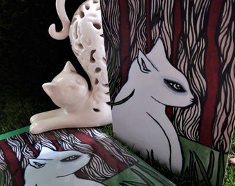 May include: A white ceramic cat figurine and two greeting cards. The cards feature a white cat illustration against a background of red and black vertical lines, resembling a forest. The figurine is a stylized cat with a long, curved tail.