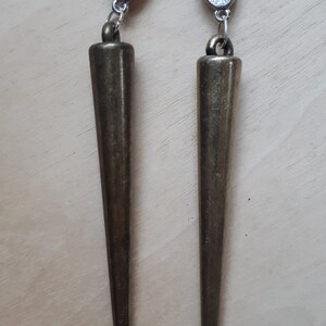 May include: A pair of bronze-colored, cone-shaped earrings. Each earring has a post back with a clear plastic disc. The earrings are suspended from a small loop. The earrings are a dark, metallic color.