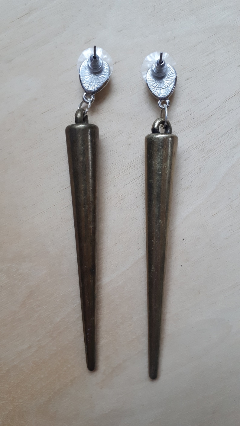 May include: A pair of bronze-colored, cone-shaped earrings. Each earring has a post back with a clear plastic disc. The earrings are suspended from a small loop. The earrings are a dark, metallic color.