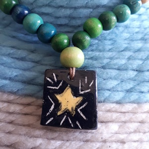 May include: A handmade necklace featuring a pendant with a yellow star design on a black background. The necklace has a string of wooden beads in shades of green, blue, and tan. The pendant is rectangular and hangs from a brown cord.