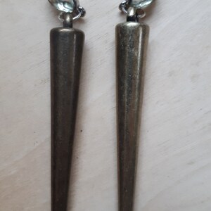 May include: A pair of long, bronze-colored earrings with a pointed, conical design. Each earring features a light green, oval-shaped stone at the top, attached to a post for pierced ears. The earrings have a vintage or antique aesthetic.