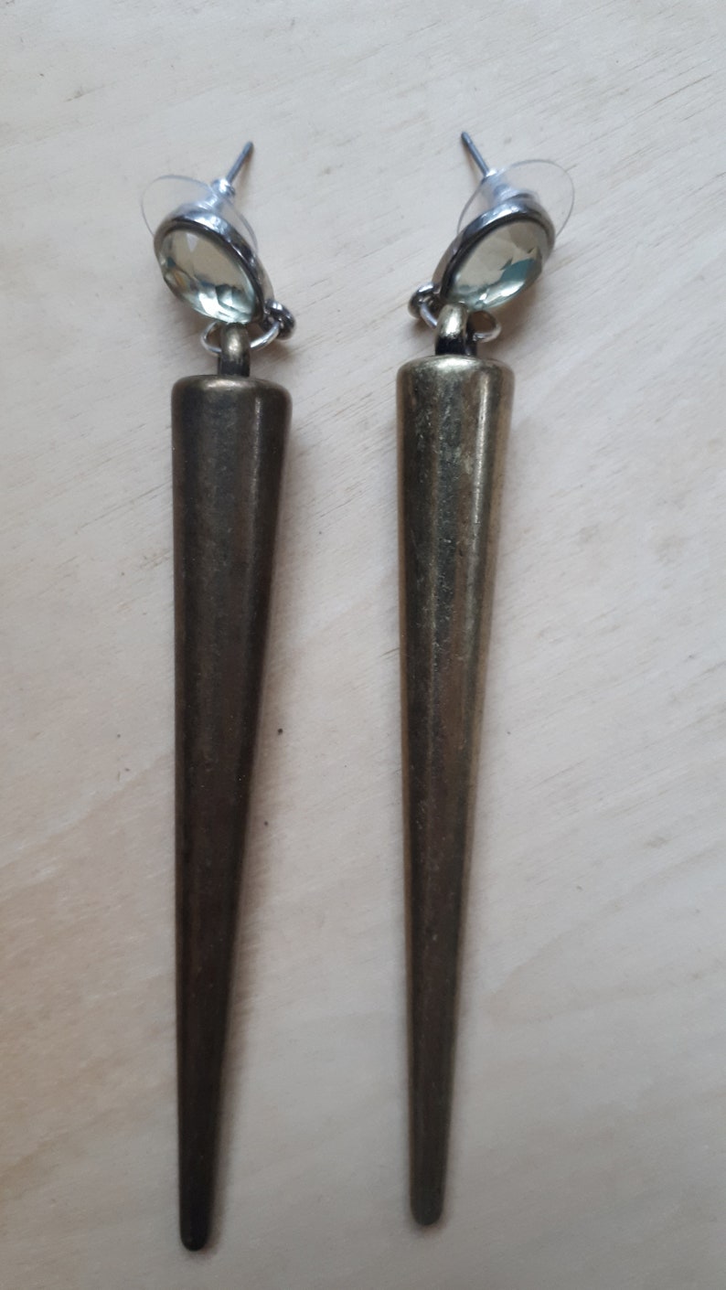 May include: A pair of long, bronze-colored earrings with a pointed, conical design. Each earring features a light green, oval-shaped stone at the top, attached to a post for pierced ears. The earrings have a vintage or antique aesthetic.