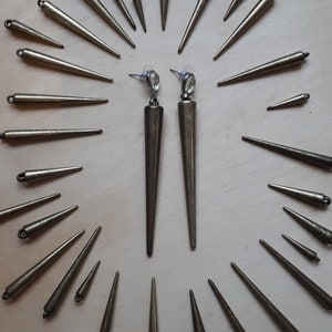 May include: A pair of long, bronze-colored earrings with a pointed, conical design. The earrings are displayed with several smaller, similar bronze spikes, creating a radial pattern. The earrings have a post backing with a clear, oval-shaped gem.