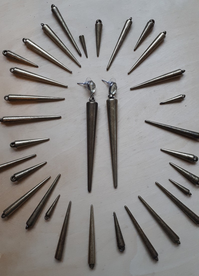 May include: A pair of long, bronze-colored earrings with a pointed, conical design. The earrings are displayed with several smaller, similar bronze spikes, creating a radial pattern. The earrings have a post backing with a clear, oval-shaped gem.