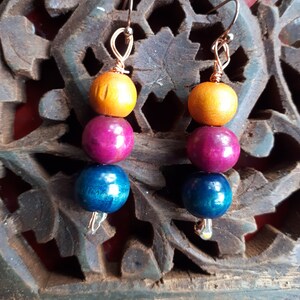 May include: A pair of handmade earrings featuring three wooden beads in orange, magenta, and blue. The beads are strung together with copper wire and have a hook closure. The earrings are displayed against a carved wooden background.