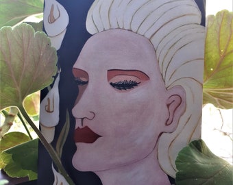 Op de afbeelding: Een schilderij van een vrouw met blond haar, rode lippen en gesloten ogen. Het kunstwerk heeft een donkere achtergrond met witte calla lelies. De vrouw draagt een parelketting. Het schilderij is omgeven door groene bladeren.