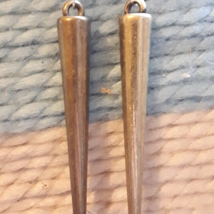 May include: A pair of earrings with a teardrop-shaped, clear crystal top and a long, bronze-colored, cone-shaped pendant. The earrings are hanging against a textured, light blue and white background.