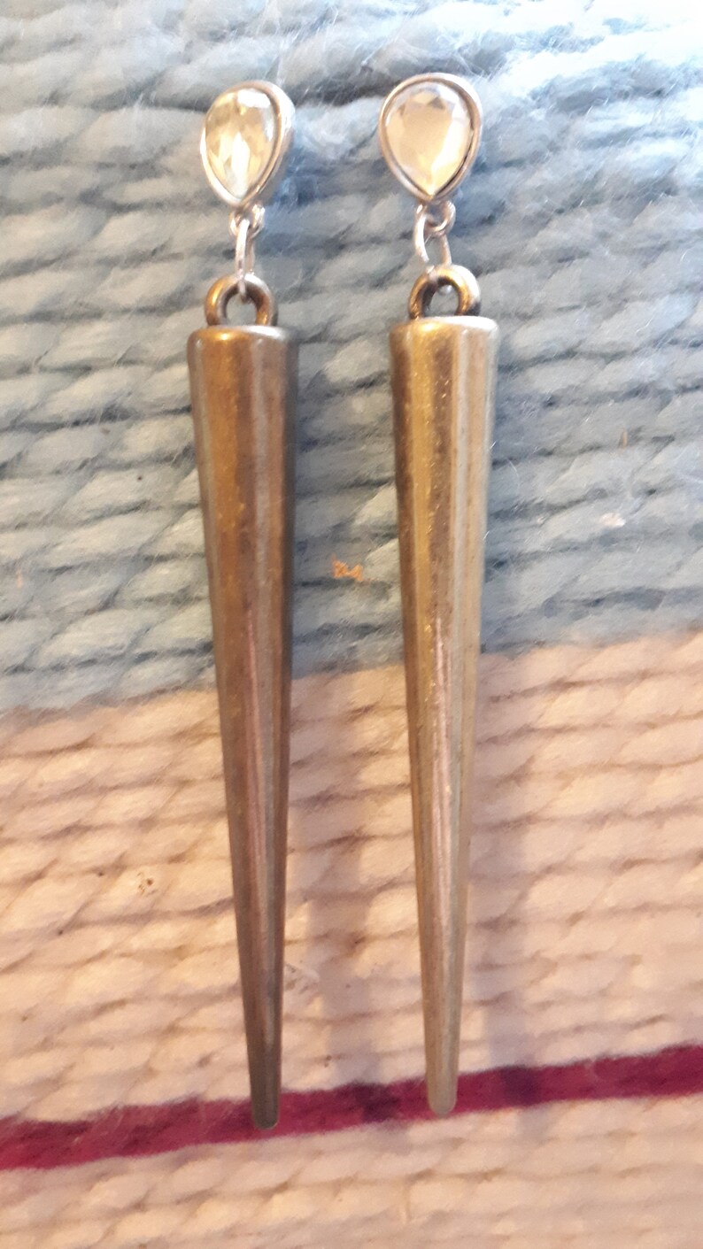May include: A pair of earrings with a teardrop-shaped, clear crystal top and a long, bronze-colored, cone-shaped pendant. The earrings are hanging against a textured, light blue and white background.