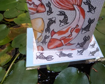 May include: A greeting card featuring a koi fish design with orange and white markings, set against a gray background with smaller black and white fish. The card is displayed amidst lily pads in water.