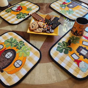 May include: Four yellow and white gingham placemats with a black border. Each placemat features a felt appliqué of a pumpkin house with a gnome peeking out the door. The pumpkin houses have brown doors and windows.