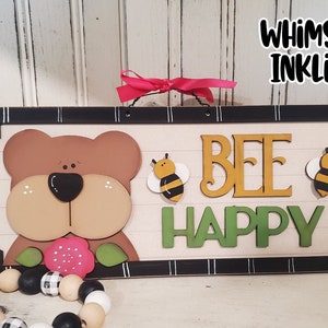 May include: Wooden sign with a whimsical design featuring a brown bear, two yellow and black bees, and the text "BEE HAPPY".