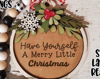 Puede incluir: Un adorno redondo de madera con el texto "Have Yourself A Merry Little Christmas" en letras negras. El adorno está decorado con hojas verdes, bayas rojas y un copo de nieve blanco. El adorno está listo para ser cortado con un cortador láser.