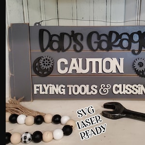 May include: A wooden sign with the text "Dad's Garage" in black lettering, "CAUTION" in white lettering, and "FLYING TOOLS & CUSSING" in white lettering. The sign is decorated with two black metal gears. The sign is on a gray background.