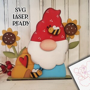 May include: A wooden gnome with a red hat, white beard, and blue pants. The gnome is holding a beehive with a red heart and a bee on each side. There are two sunflowers and a bee on the gnome's hat. The image includes the text "SVG LASER READY".