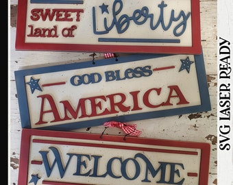 Może przedstawiać: Cztery drewniane tabliczki pomalowane na czerwono, biało i niebiesko. Tabliczki przedstawiają napisy "Sweet land of Liberty", "God Bless America", "God Bless America" i "Welcome".
