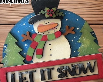 May include: A wooden snowman ornament with a red and green scarf and a black hat. The ornament is painted with a blue background and white snowflakes. The ornament has a sign that says "Let It Snow".