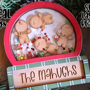 Op de afbeelding: Een kerstversiering met een rood en groen frame, met peperkoeken poppetjes met namen in een heldere bol. De basis luidt "The Mahughs". De versiering is versierd met kleurrijke confetti en de tekst "Whimsical Inklings" en "SVG Laser Ready".